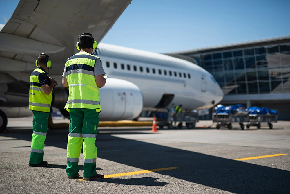 Airport crew in Atire brand high-vis gear stand on a tarmac near a large commercial plane.