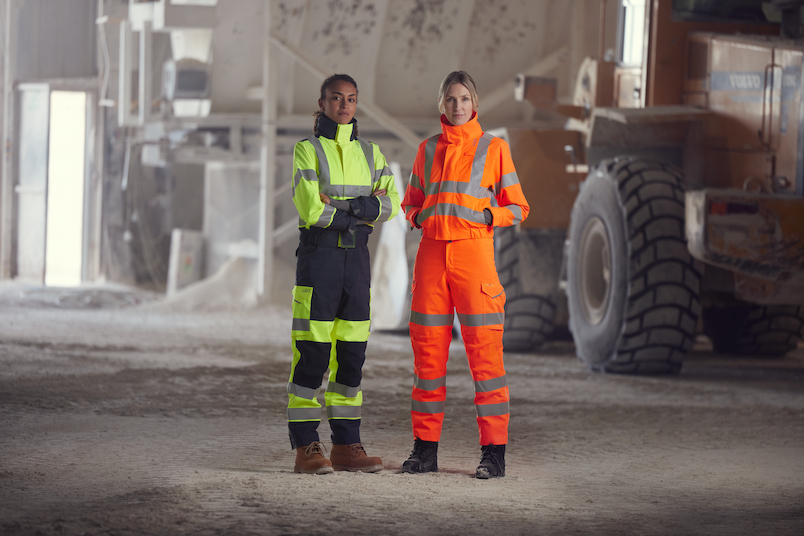 Two women wearing Atire high-visibility construction workwear in an industrial setting with heavy machinery.
