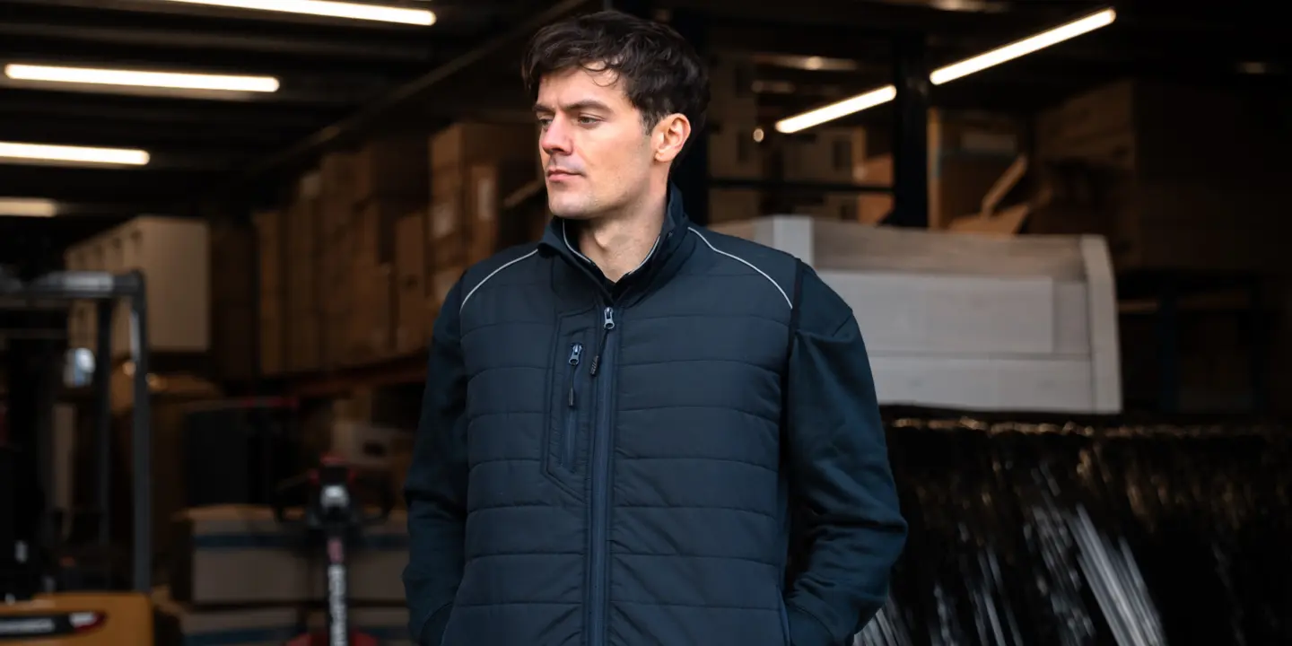 A man in an Atire brand navy quilted gilet stands in a warehouse with storage boxes and a forklift