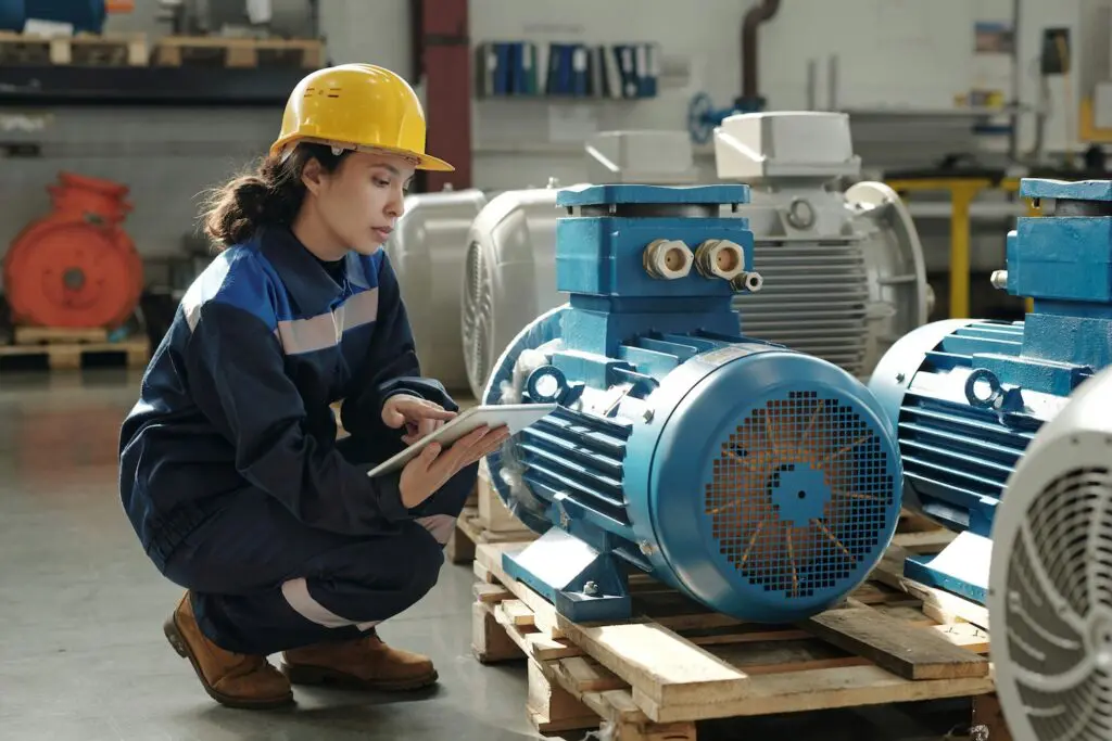 Female engineer wearing Atire industrial workwear: navy coveralls and yellow hard hat in a factory setting