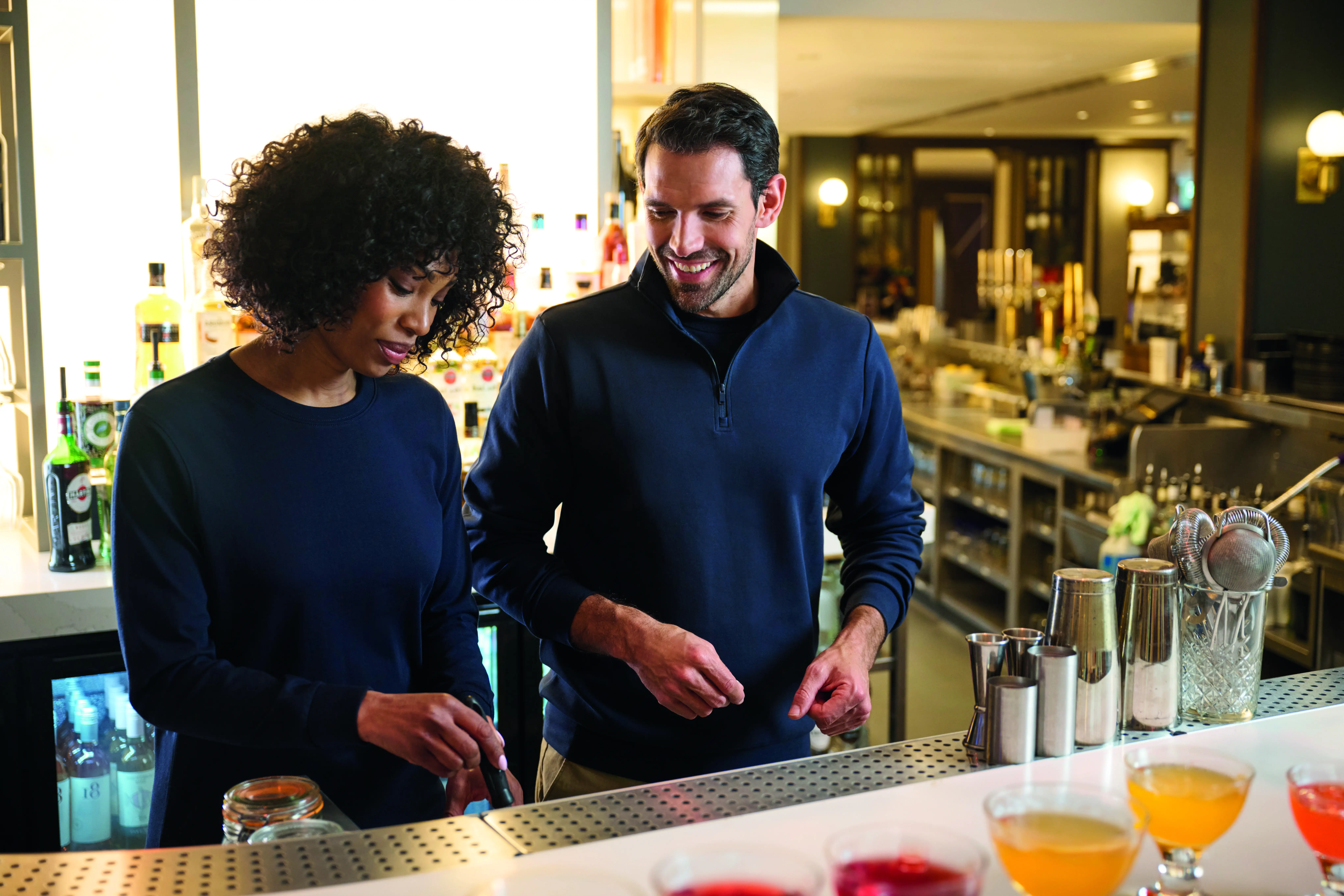 Bar staff in a navy sweatshirt and quarter zip top preparing cocktails behind a restaurant bar