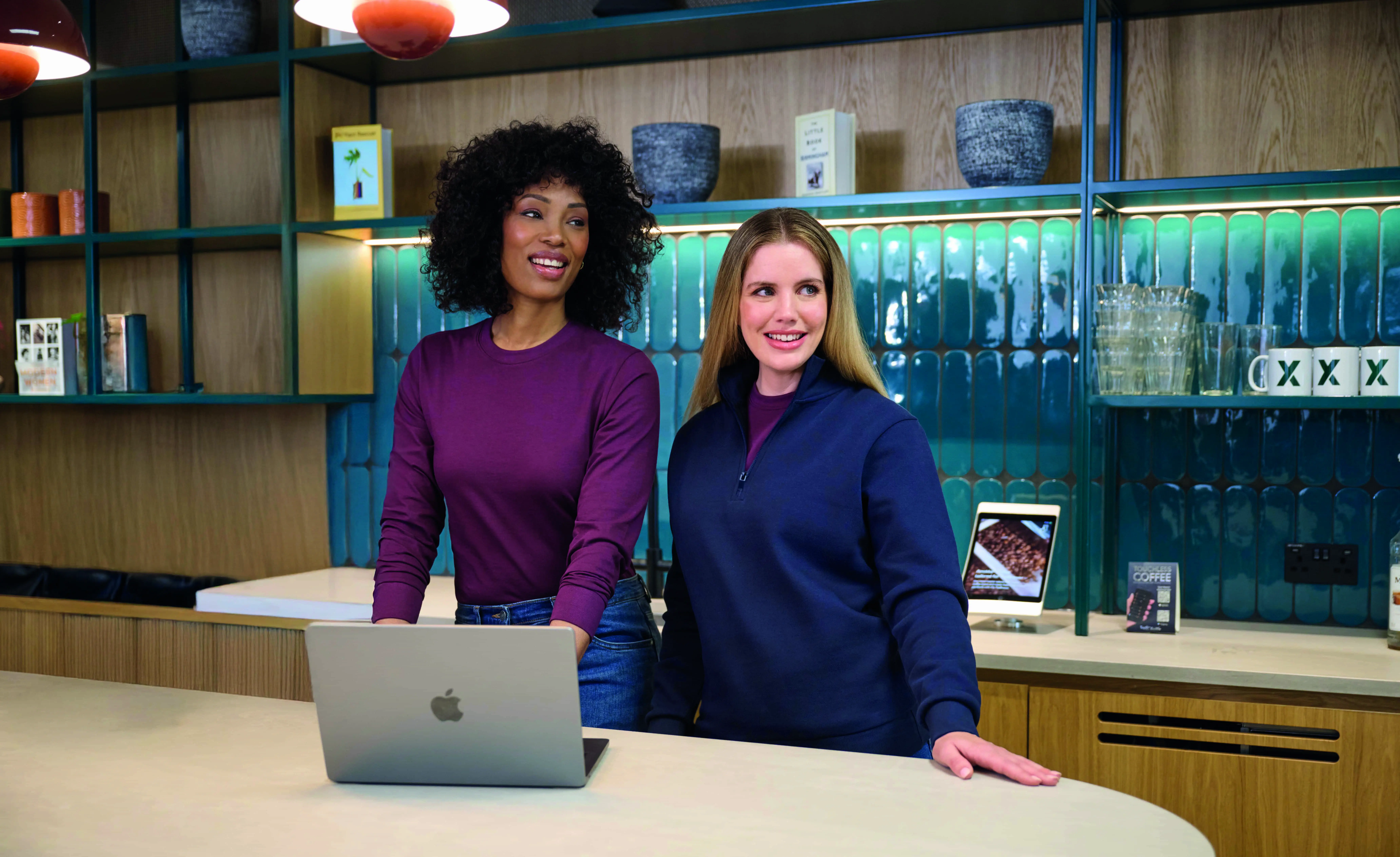 Hotel reception staff in a plum sweatshirt and navy quarter zip top at a front desk with a laptop