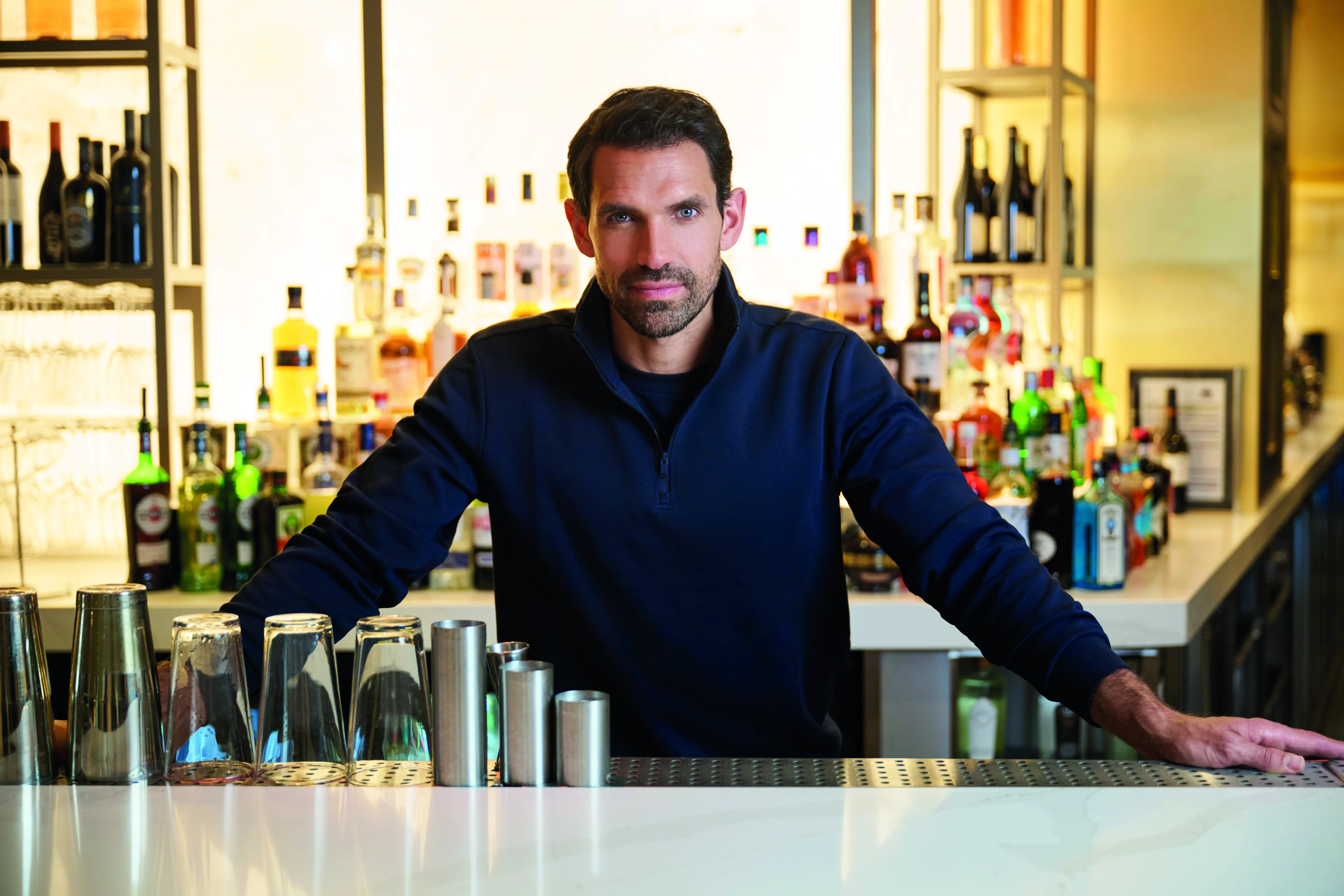 Bartender in a navy quarter zip top standing behind a bar with glasses, shakers and spirits on display