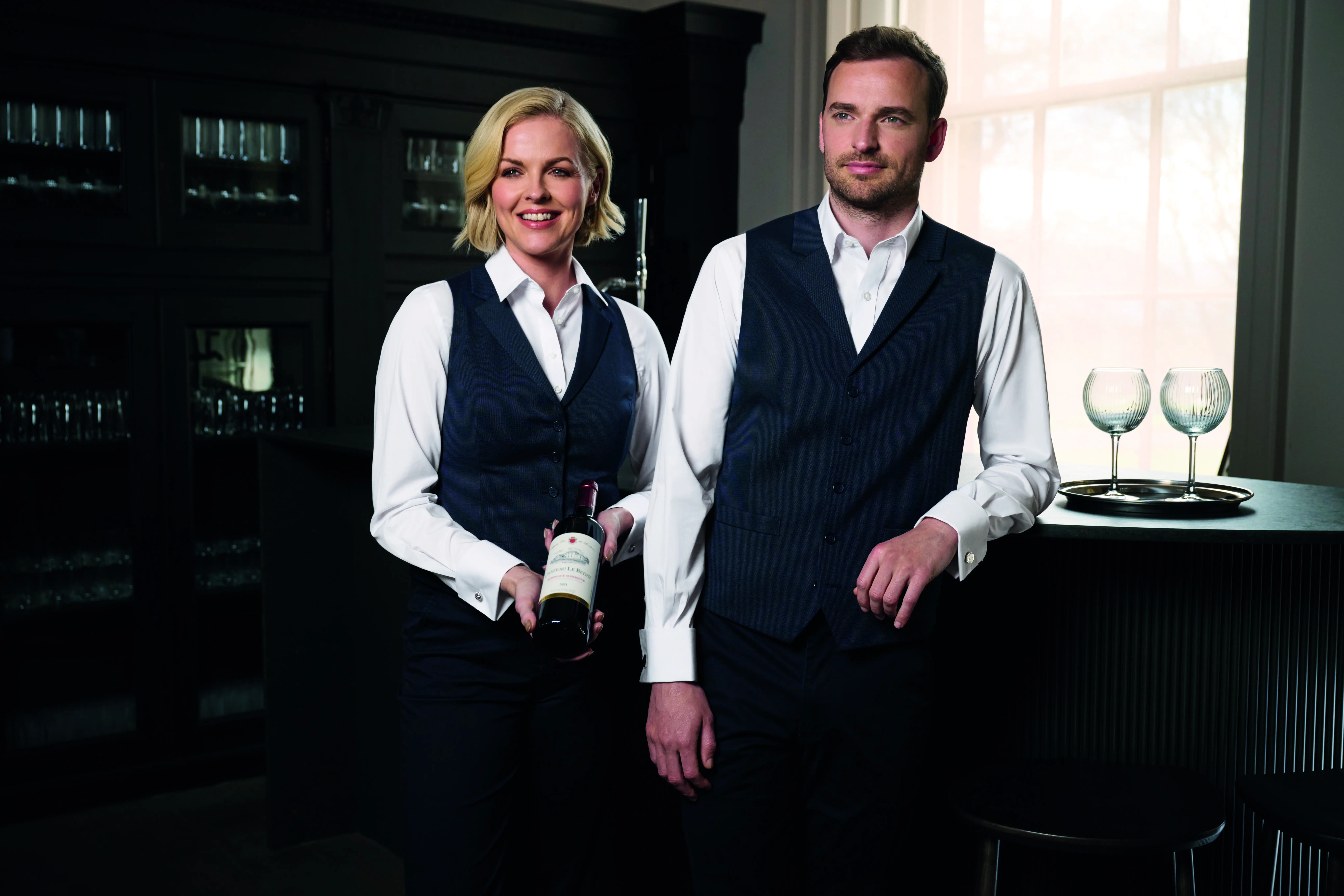 Hotel bar staff in navy waistcoats and white shirts, one holding a bottle of wine beside a tray of glasses