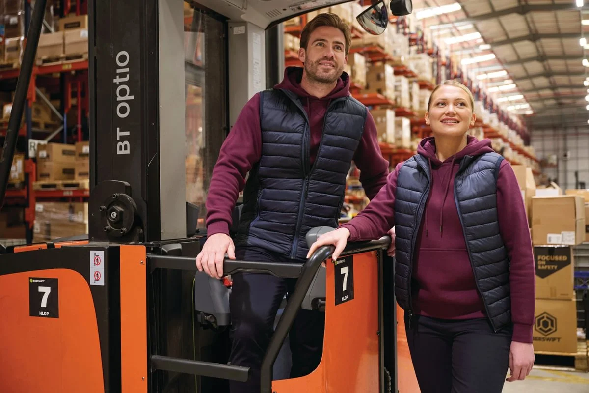 Warehouse staff in navy quilted bodywarmers and burgundy hoodies beside a forklift truck in racking aisles